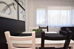 Ein Essbereich mit einem weißen Tisch und schwarzen Stühlen, ein schwarzes Kissen auf dem Tisch sowie ein Fenster mit leicht transparenten Vorhängen.
