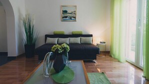 Una sala de estar con un sofá negro, cojines verdes, un cuadro en la pared, una planta en maceta y un florero con flores.