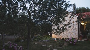 A stone building with a red-tiled roof, surrounded by a garden with purple flowers and a tree.