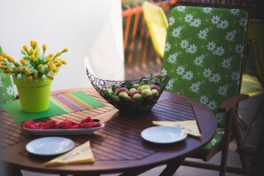 Ein runder Holztisch mit einem grünem, blumengemusterten Stuhl, einem Blumentopf, einer Obstschale und einer Tellervoll Wassermelone.