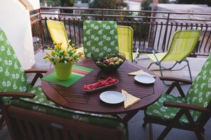 Ein runder Holztisch mit grün karierten Stühlen, ein Blumentopf und eine Obstschale auf einem Balkon.