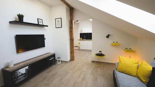 A modern living room with a flat-screen TV, a wooden console table, a sofa with yellow cushions, and a kitchen in the background.