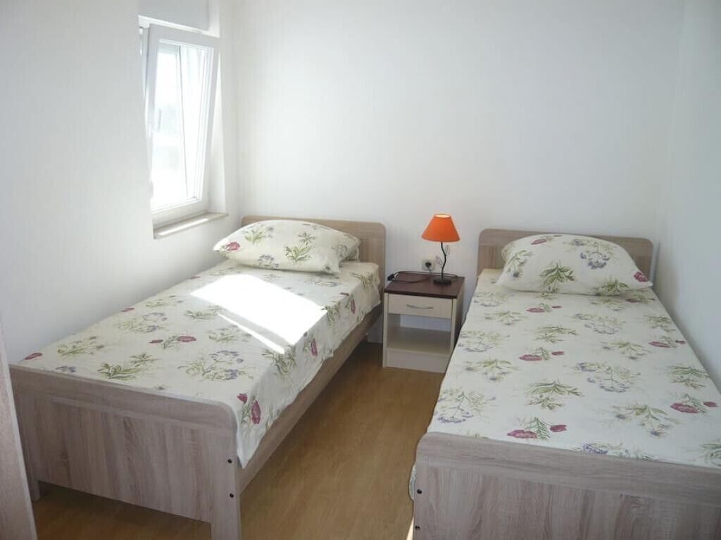 Une petite chambre propre, avec deux lits simples, une table de chevet, une lampe et une fenêtre.