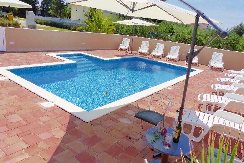 A rectangular swimming pool with a white border, surrounded by a tiled patio. There are white lounge chairs and a table with a bottle and a cup on it.