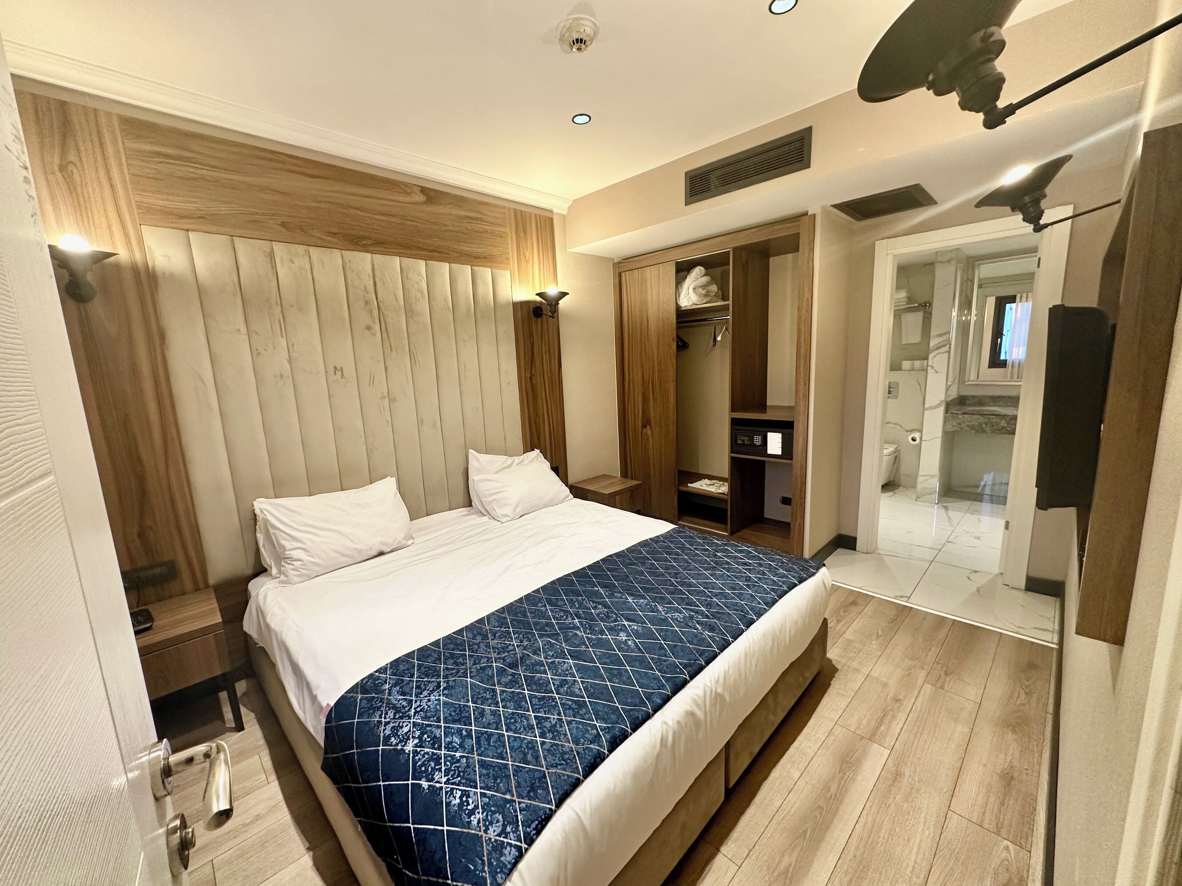A modern hotel room with a large bed, wooden headboard, built-in closet, and a walk-in shower.