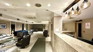 Um hall de hotel com balcão de recepção, área de estar e corredor que dá acesso a outros quartos.