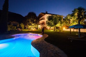 A swimming pool with a stone edge, surrounded by a well-lit garden area with trees and a building in the background.