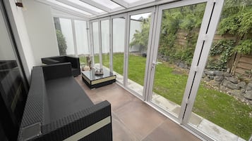 Un espace patio moderne avec un canapé en osier noir et une table basse en verre, ainsi que de grandes portes-fenêtres coulissantes donnant sur un jardin.