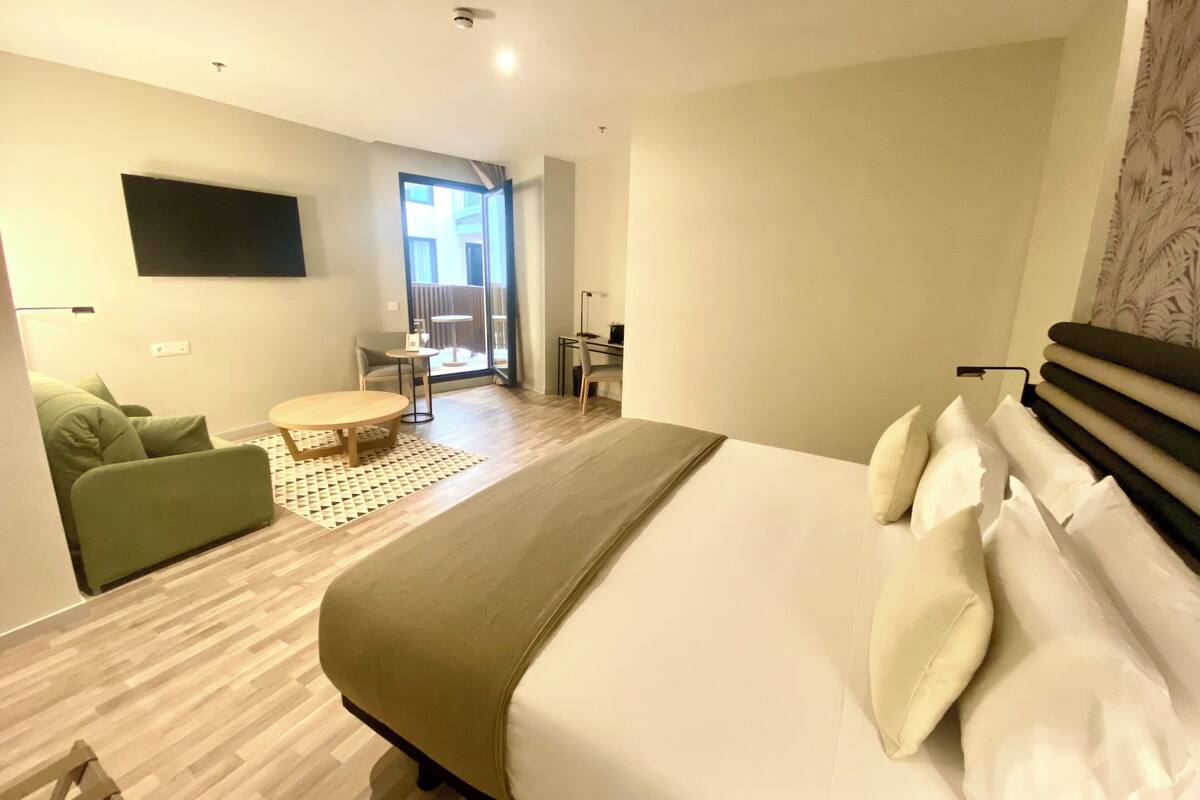 A modern hotel room with a large bed, a green sofa, a small round table, and a balcony with a view.