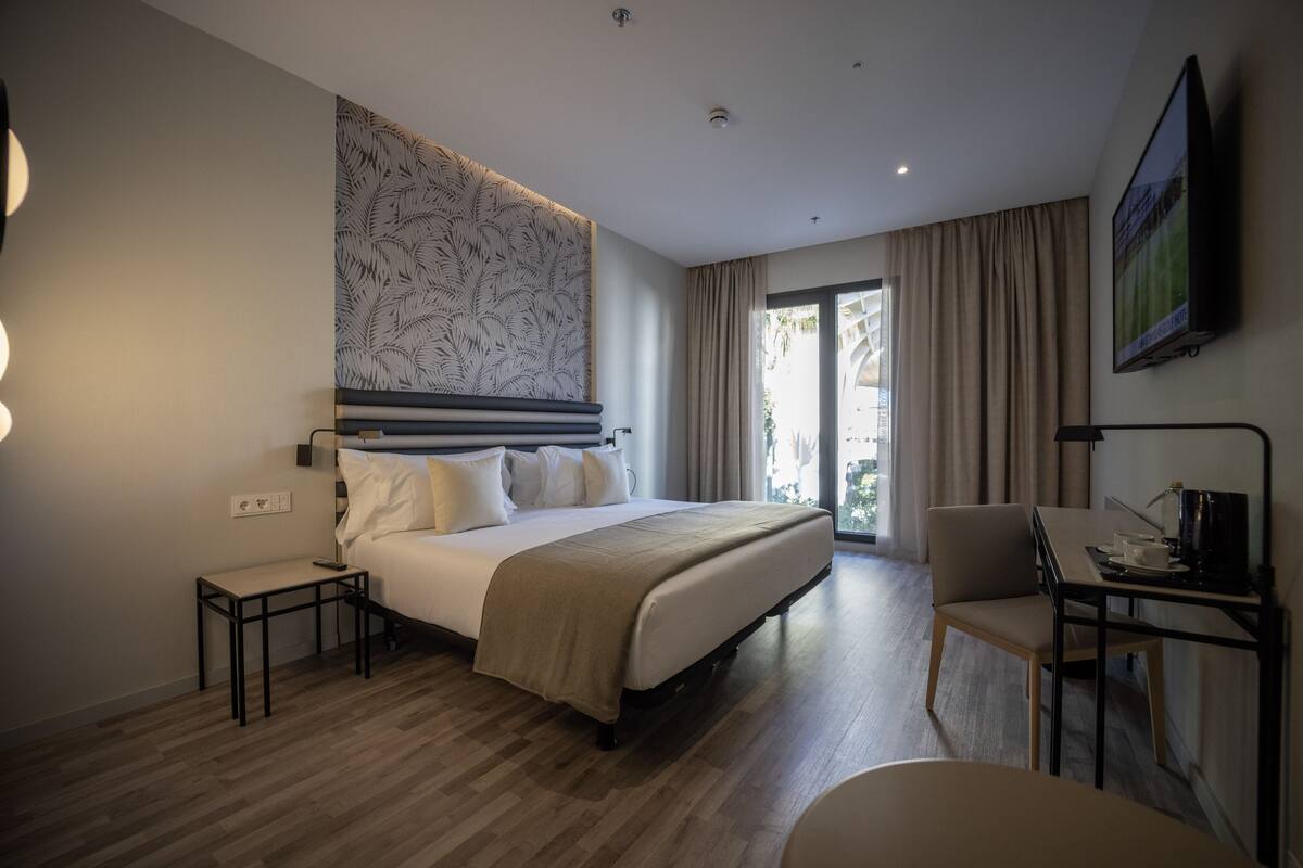 A modern hotel room with a large bed, a desk with a chair, a TV, and a view of the outdoors.
