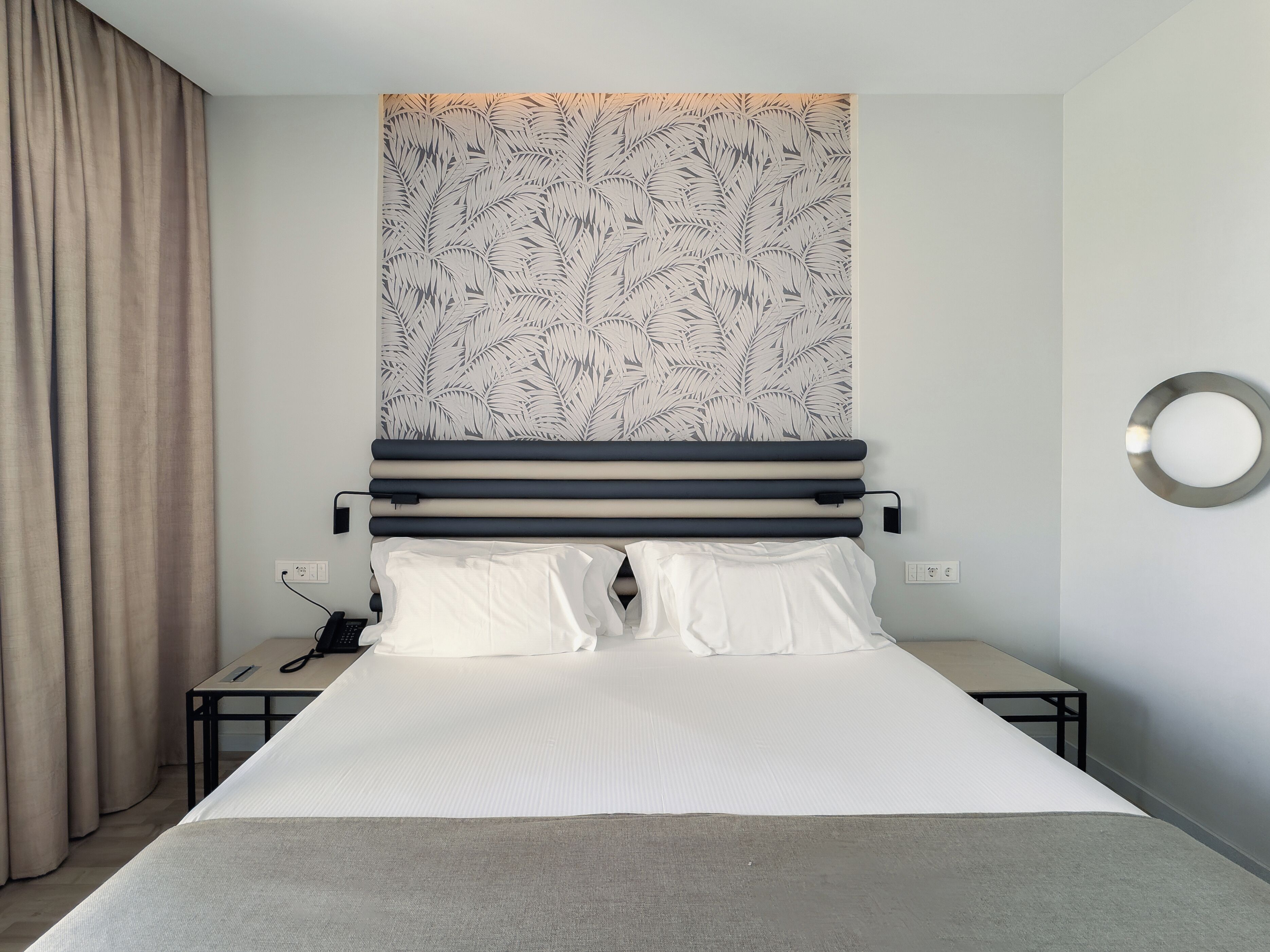 A neatly made bed with white linens, a headboard with a leaf pattern, a bedside table with a phone, and a wall-mounted light fixture.