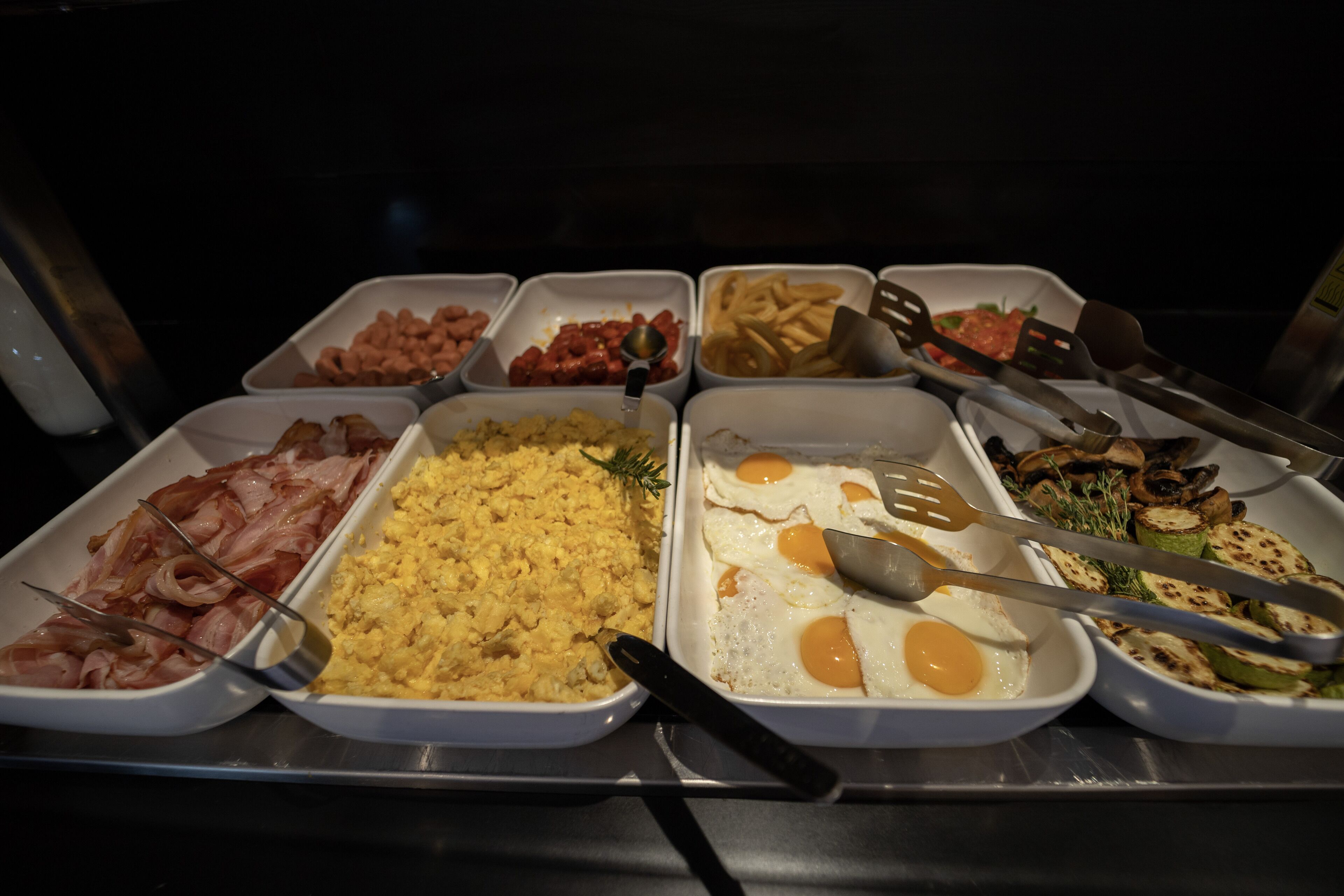 Un bufé con distintos alimentos, incluyendo tocino, huevos revueltos y verduras.
