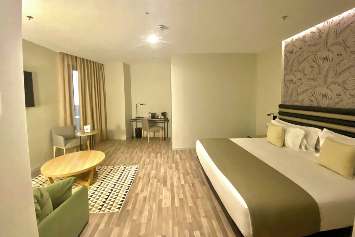 A modern hotel room with a large bed, a desk with a chair, a small table, and a patterned headboard.