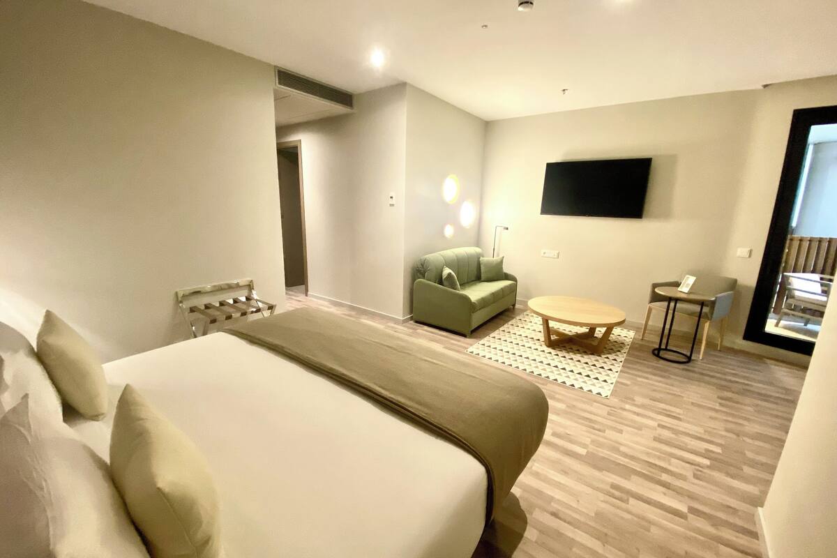 A hotel room with a large bed, a green sofa, a small round table, and a TV mounted on the wall.