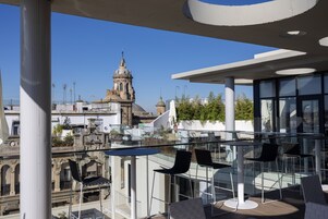 Una terraza en la azotea con mesas y sillas de vidrio, que ofrece vistas de edificios históricos y un cielo azul despejado.