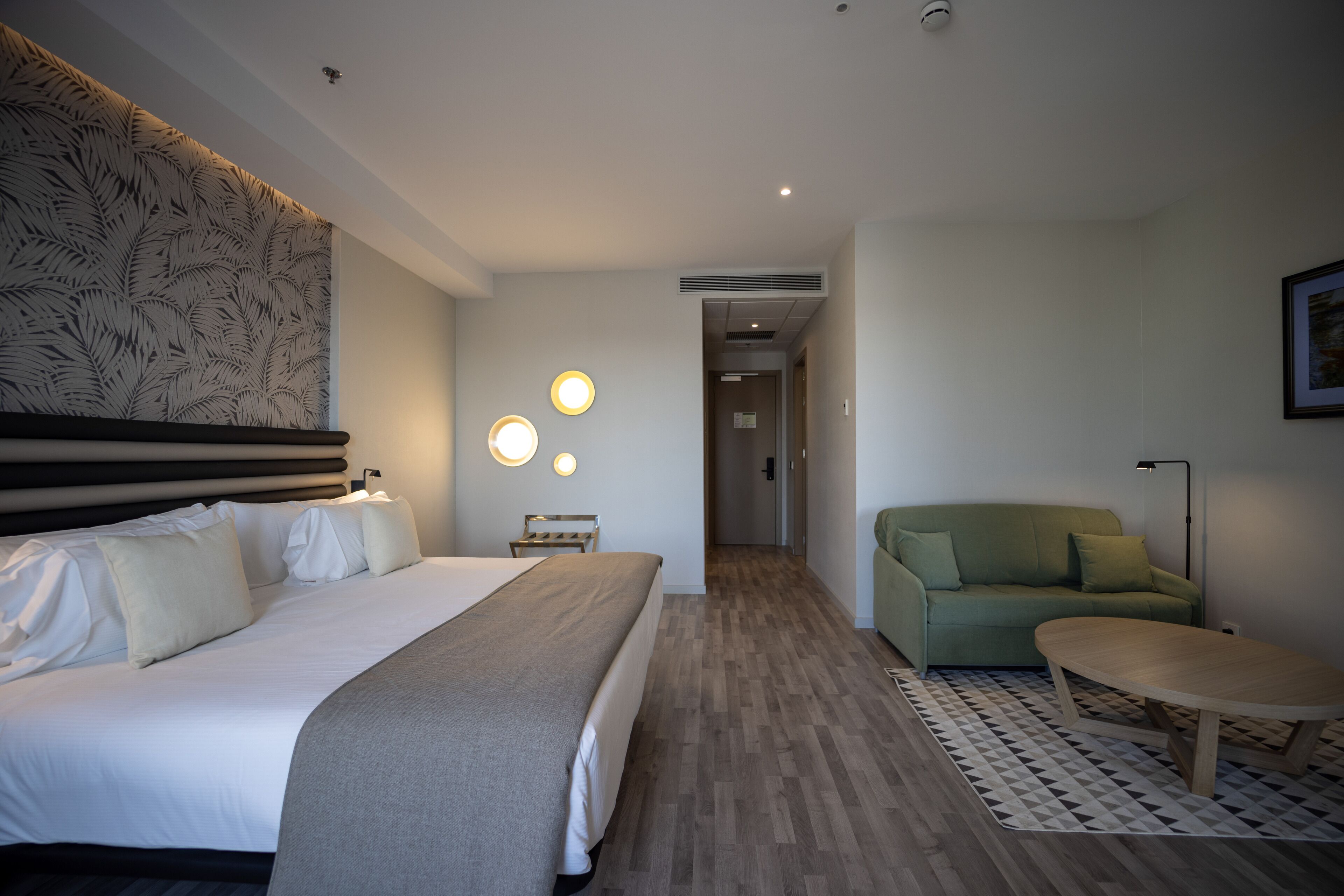 Una habitación de hotel con una cama grande, un sofá verde, una mesita pequeña y una alfombra estampada.