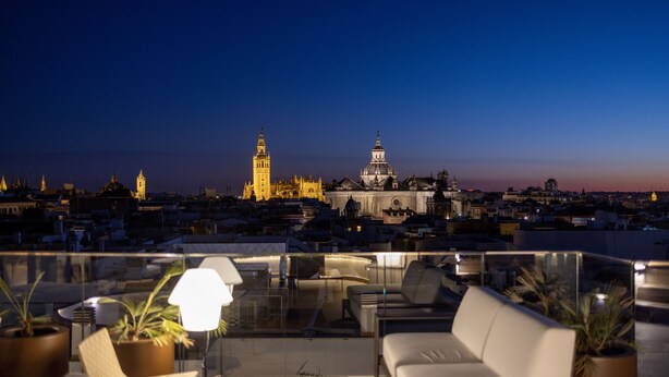 Terraza en la azotea con vistas a edificios históricos, incluyendo una torre y una cúpula, amueblada con un sofá, sillas y una mesa.