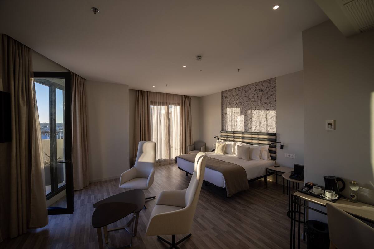 A modern hotel room with a bed, two armchairs, a small table, and a coffee station.