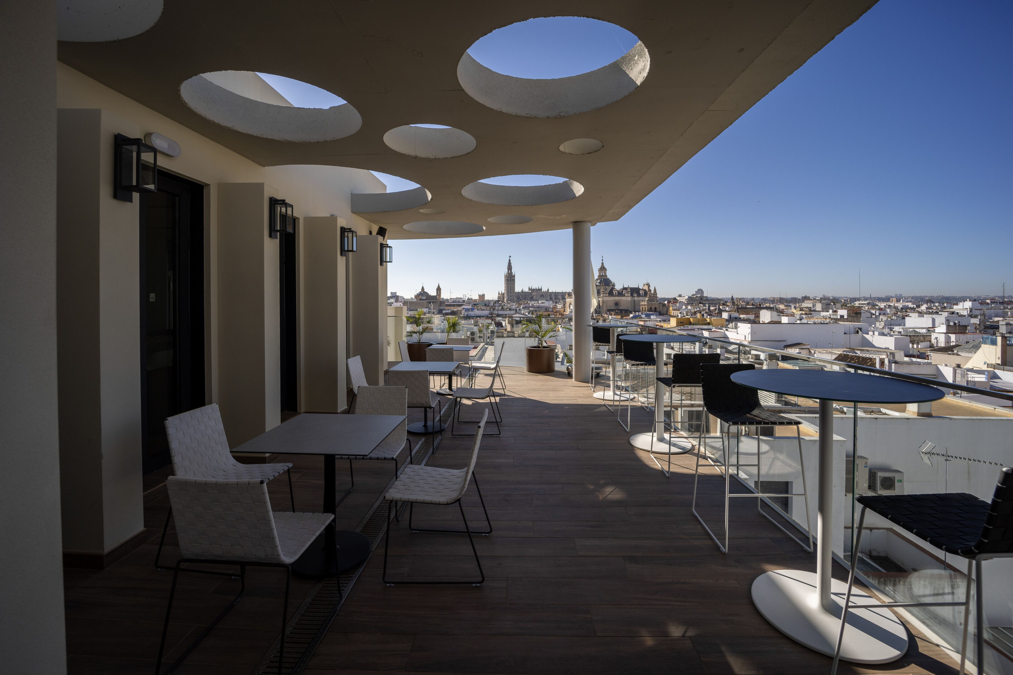 Eine Terrassenanlage im Freien mit Tischen, Stühlen und einem besonderen Dachdesign, mit Blick auf die Stadt.