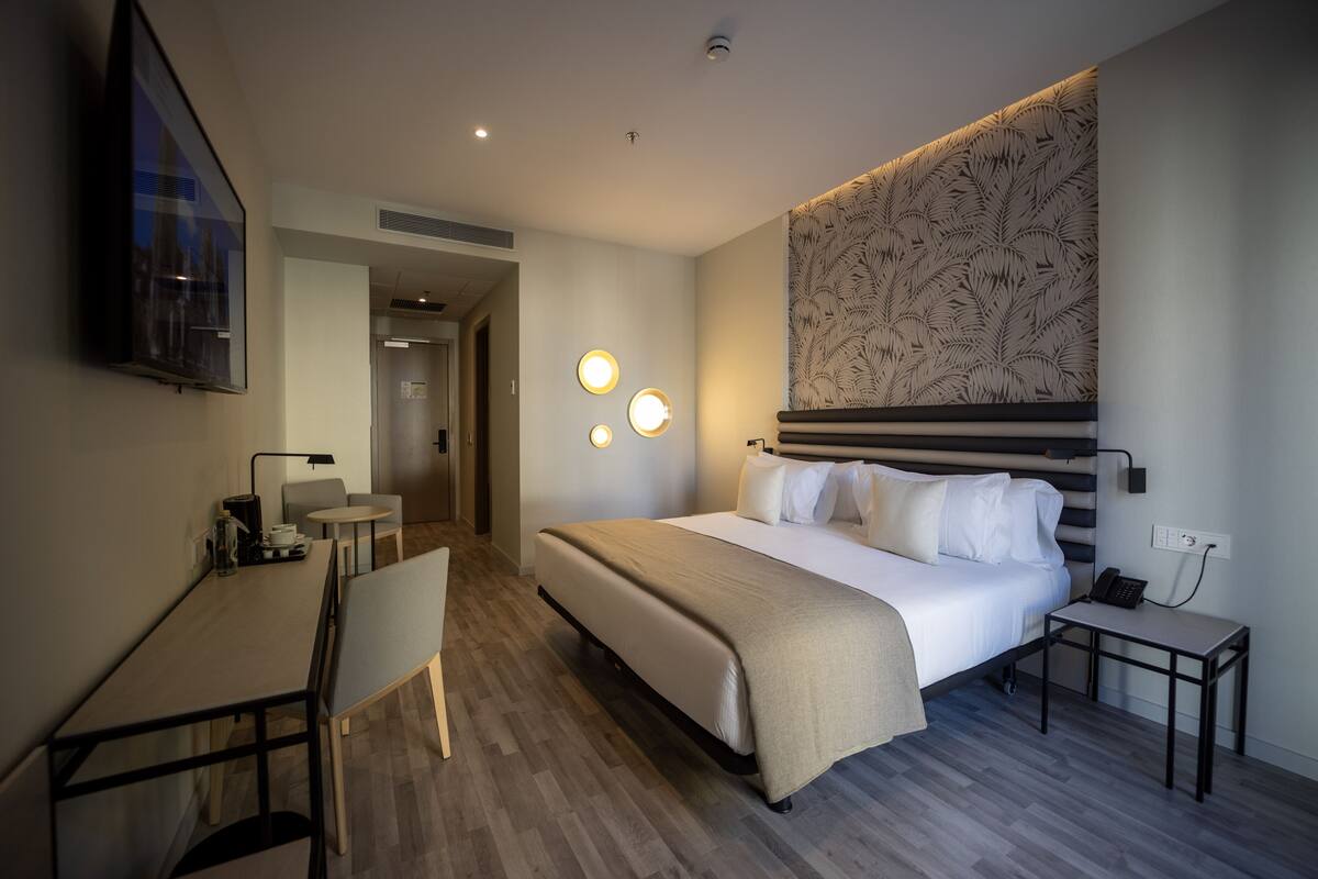 A modern hotel room with a large bed, a desk, a chair, and a small table. There are wall sconces and a ceiling light.