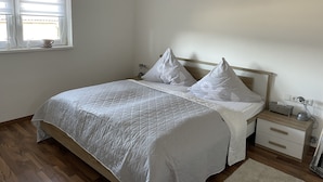 A bedroom with a wooden bed, white bedding, a bedside table with a lamp, and a window with blinds. - Ferienwohnung im Pfalz (Lustadt)