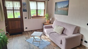 Una sala de estar con puerta de madera, ventana con cortinas, sofá rosa, una mesita, una lámpara y un cuadro en la pared.