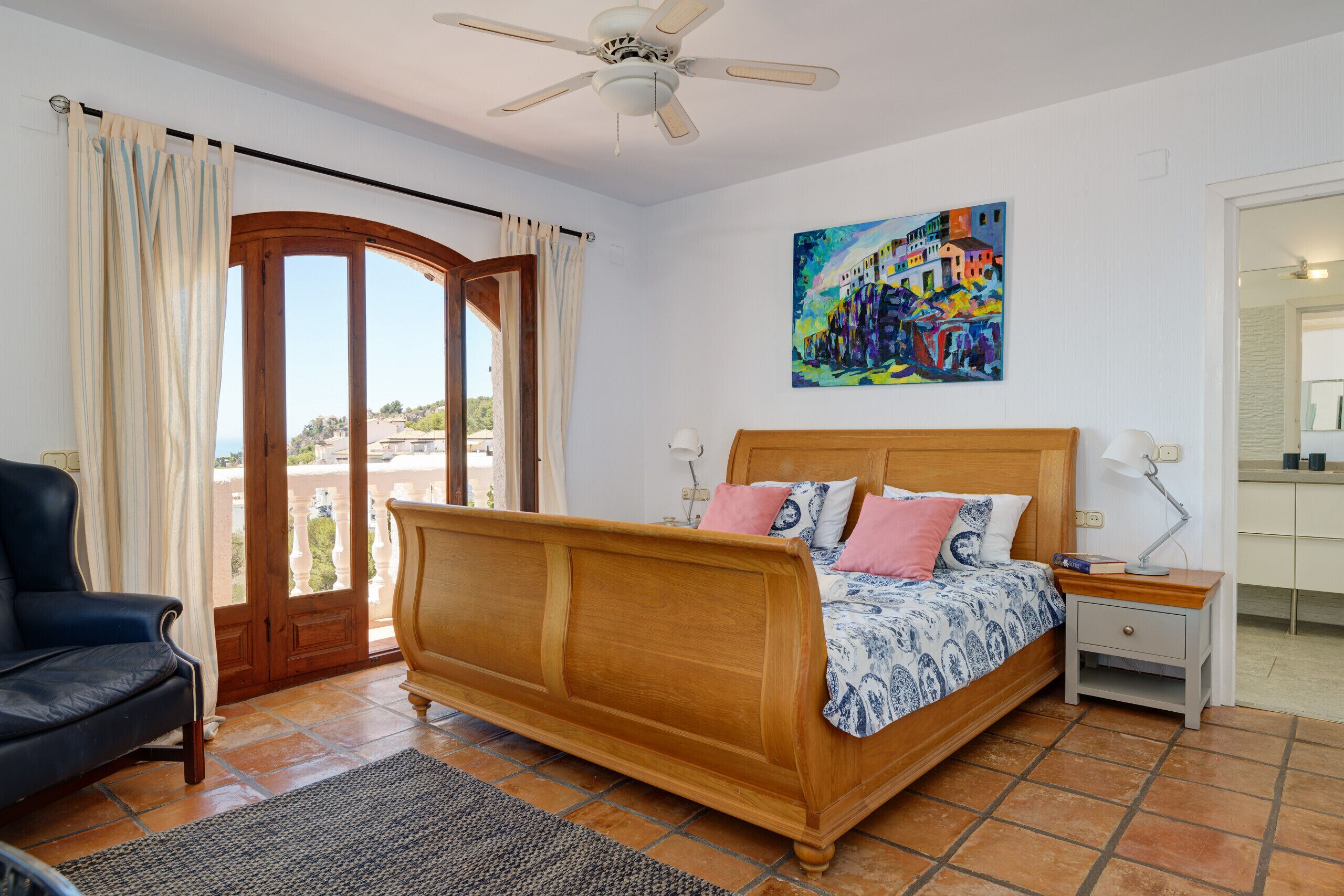 A bedroom with a wooden bed, a nightstand, a chair, a ceiling fan, and a painting on the wall.