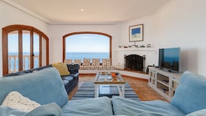 A living room with a sofa, coffee table, and TV, a fireplace, and a view of the sea.