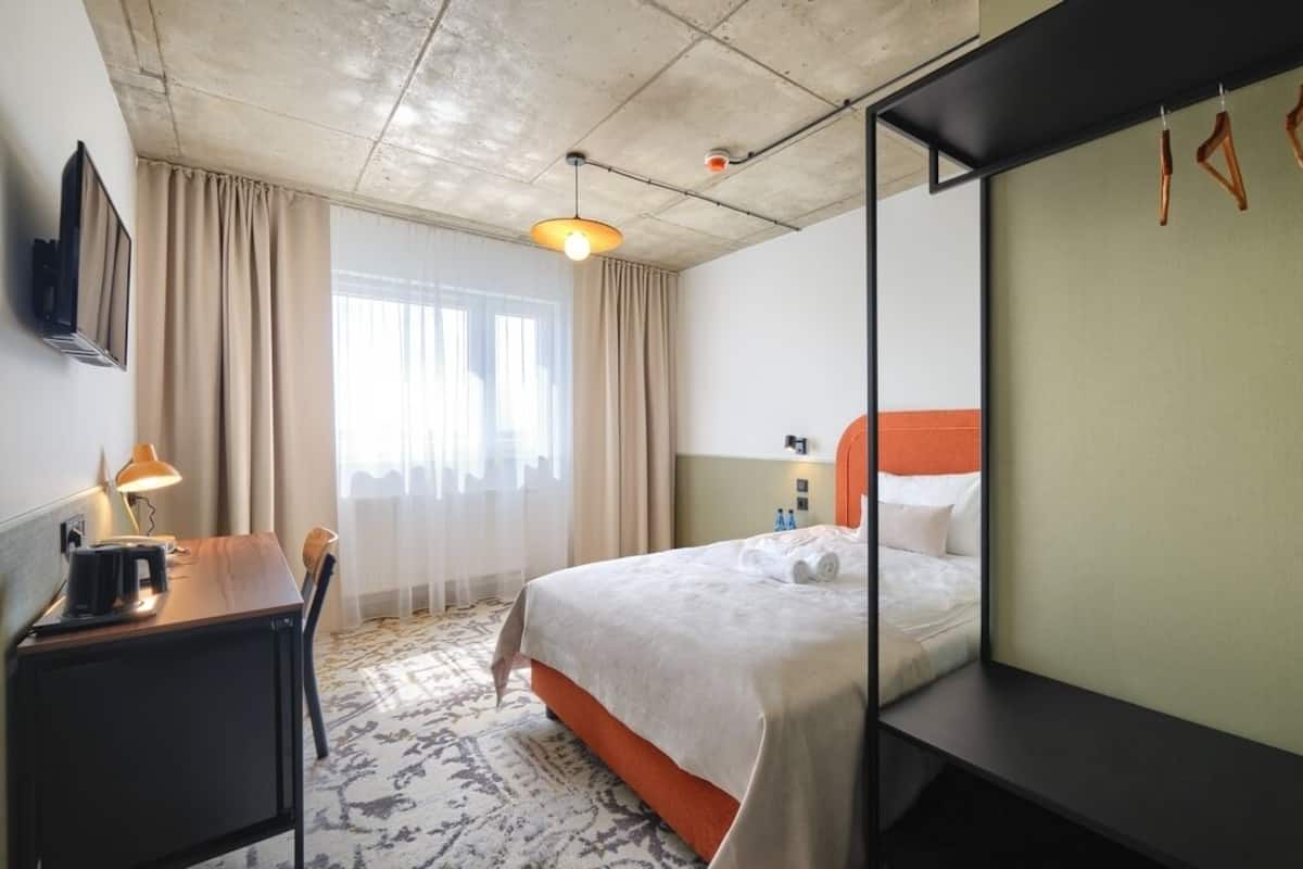 A hotel room with a bed, a desk, a chair, a lamp, a TV, and a window with curtains.
