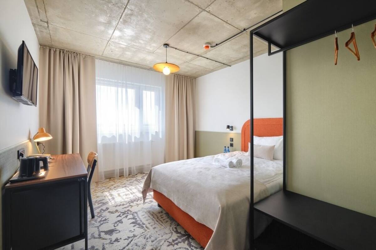 Habitación de hotel con cama, escritorio, silla, lámpara, televisor y ventana con cortinas.
