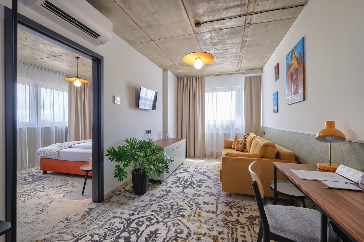 A hotel room with a bed, a sofa, a desk with a lamp, a potted plant, and a TV.