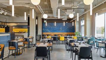 Um restaurante moderno com pé-direito alto, iluminação pendente e uma parede de mármore. Mesas dispostas para refeições, e uma área de bar visível ao fundo.