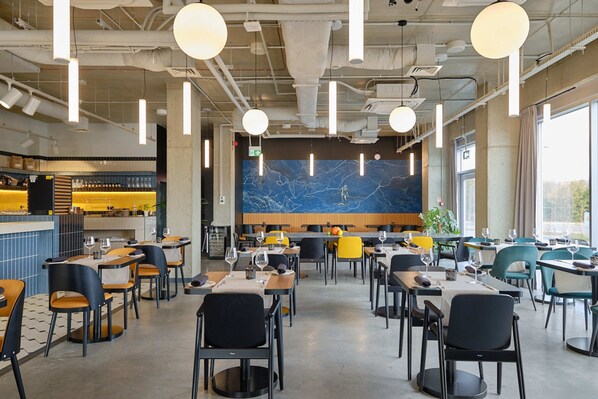 A modern restaurant with high ceilings, pendant lighting, and a marble wall. Tables are arranged for dining, and there is a bar area visible in the background. - Arche Hotel Wrocław Airport (Wroclaw)