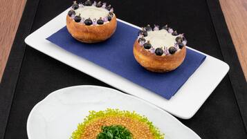 Een wit bord met een gerecht, versierd met groene kruiden en gele stipjes, en een rechthoekig bord met twee gerechten, belegd met bosbessen en paarse bloemen.