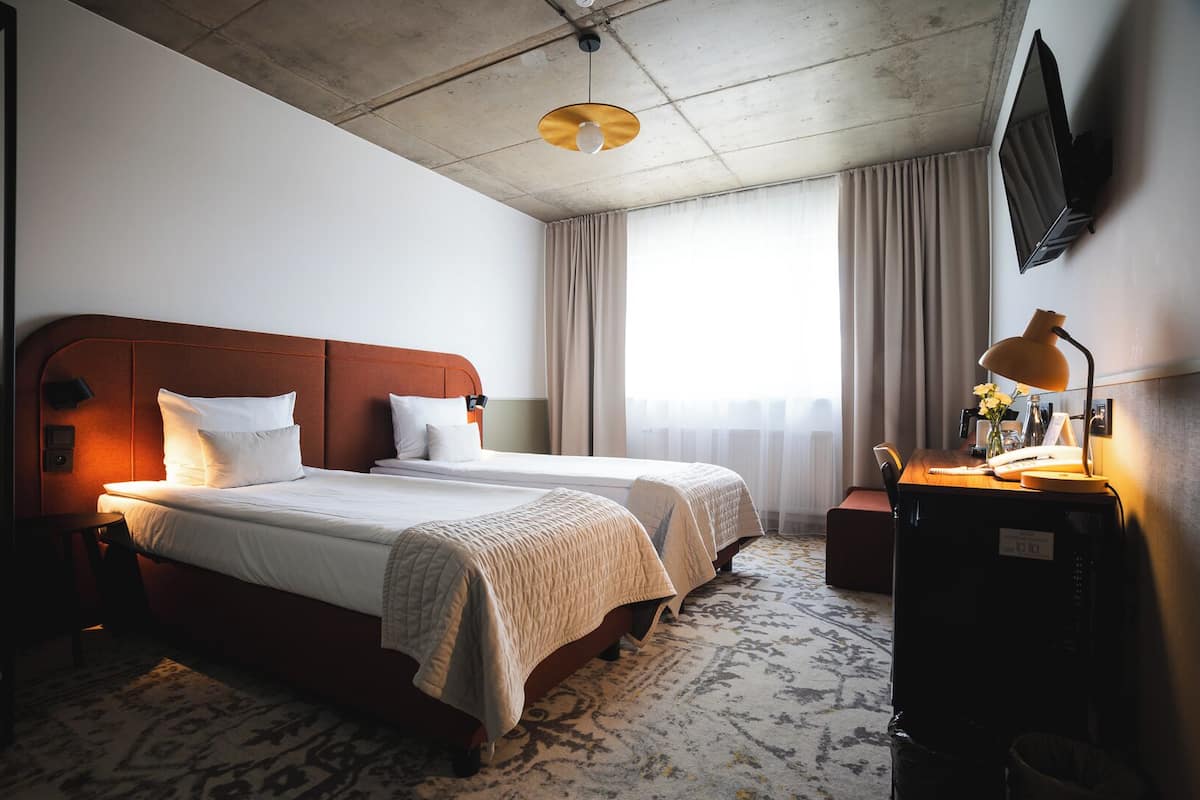 A hotel room with two beds, a desk with a lamp, a TV mounted on the wall, and a large window with curtains.