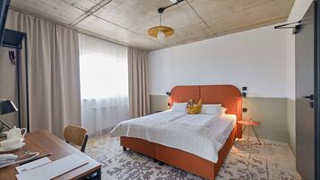 A hotel room with a bed, a desk with a chair, a small table, and a window with curtains.
