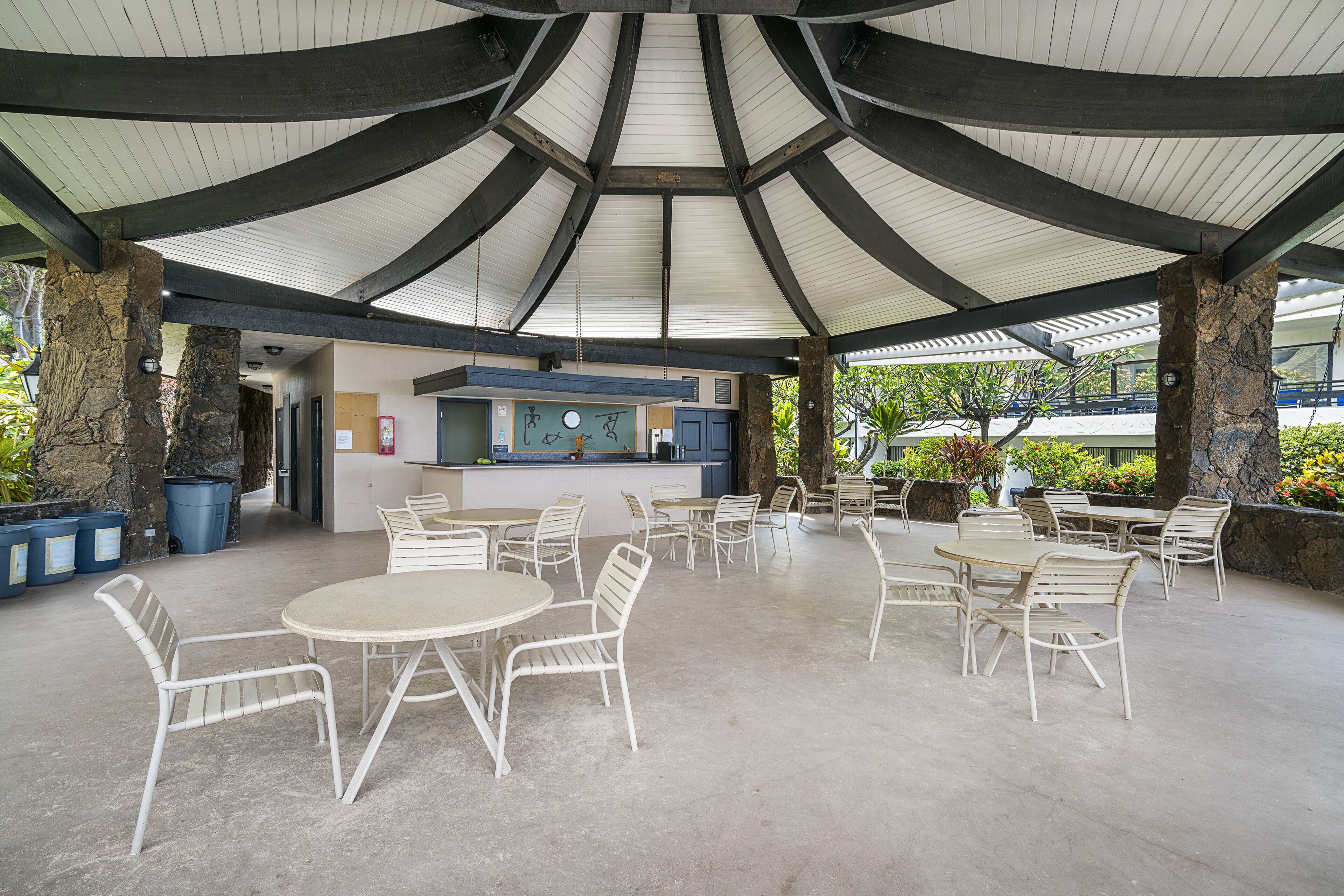 An outdoor patio area with round tables and white chairs, a stone pillar, and a covered seating area with a view of the surrounding greenery.