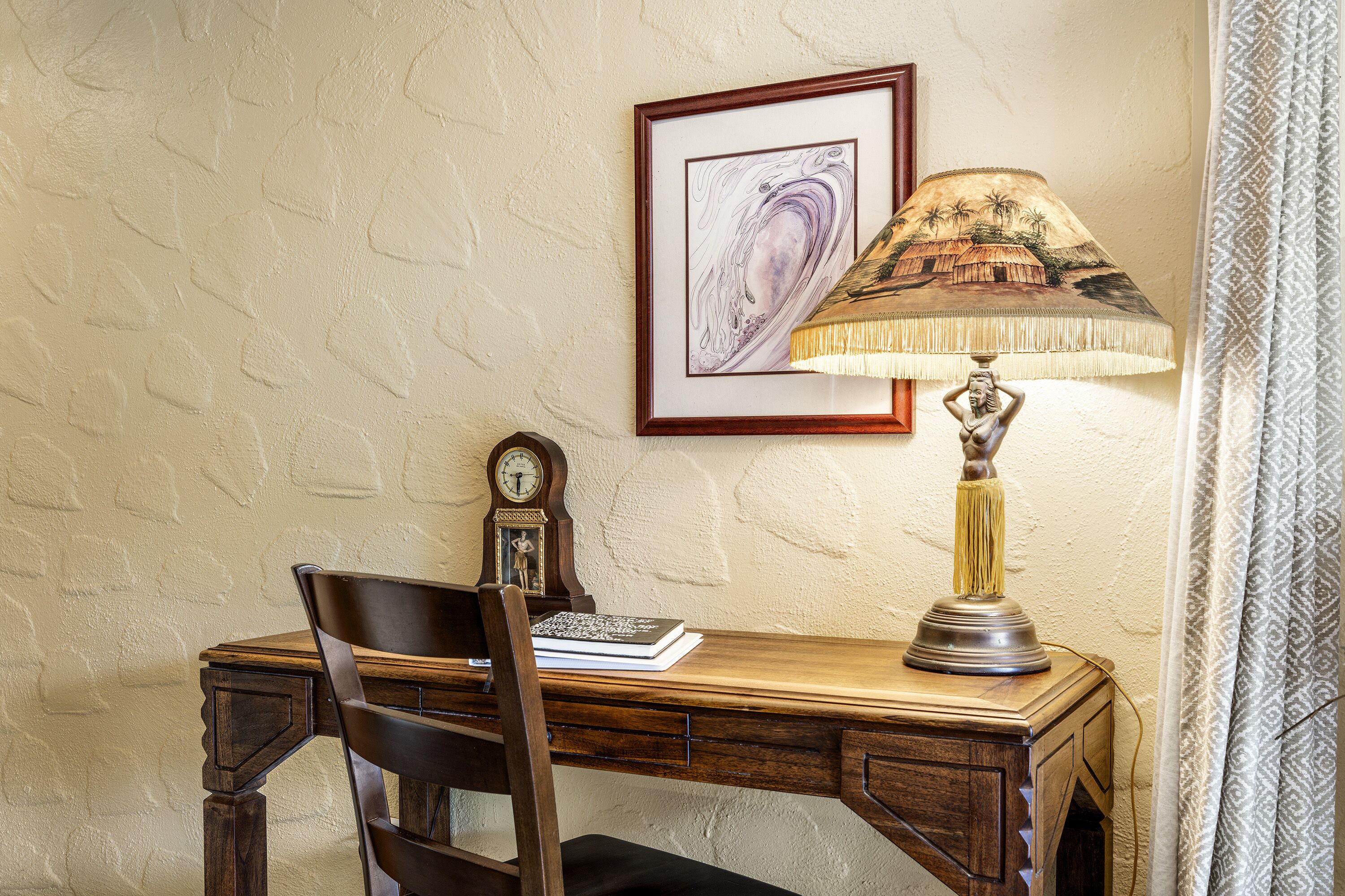 Un bureau en bois avec une chaise, un tableau encadré au mur, une lampe et une horloge.