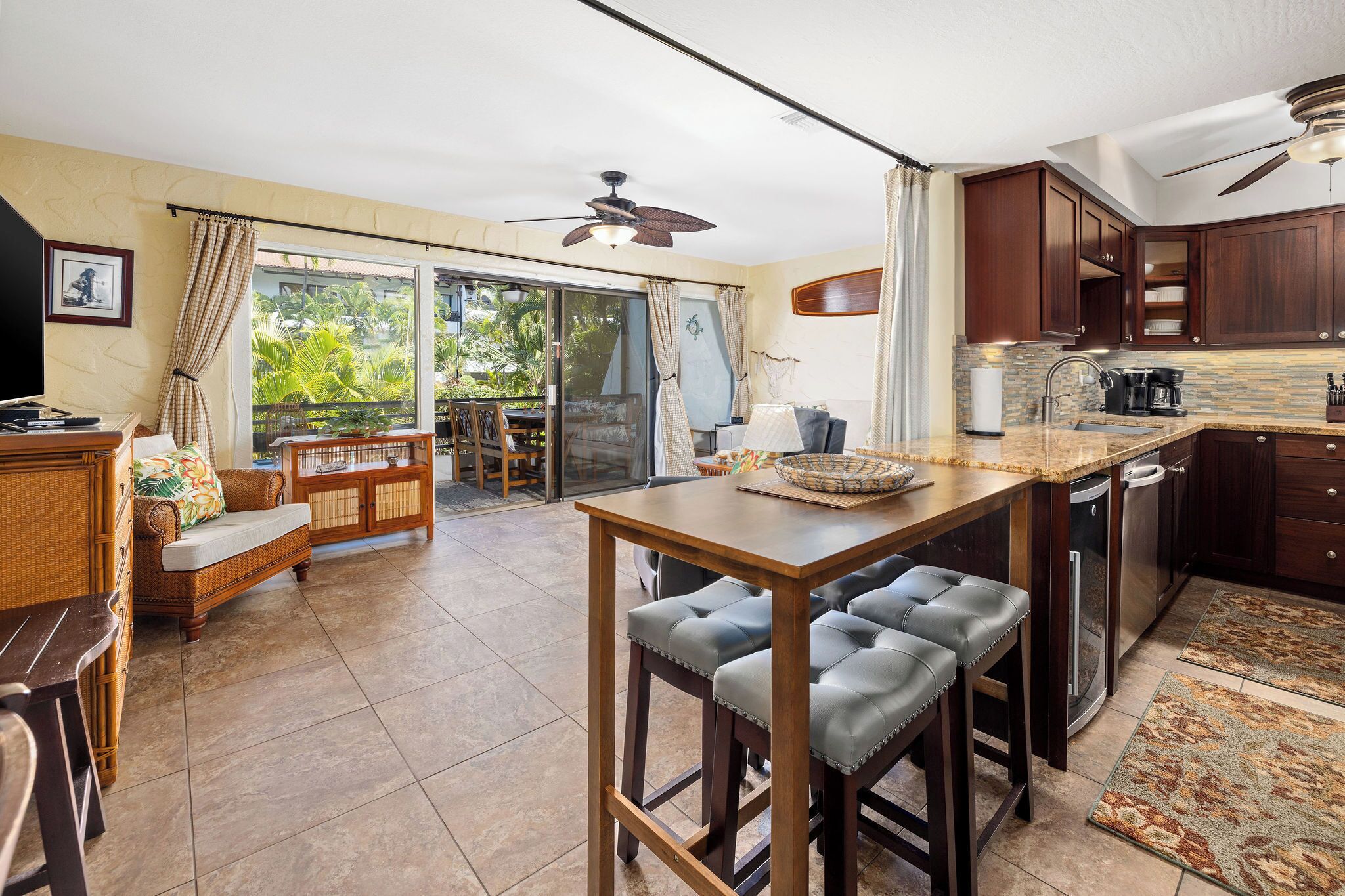 Un'ampia cucina con mobili in legno scuro, un'isola centrale con sgabelli, una zona pranzo con divanetto e tavolo in vimini e una porta scorrevole in vetro che dà su un balcone con vista sul verde.