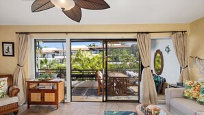 A living room with a sofa, a wooden coffee table, and a glass table with a decorative item. There is a sliding glass door leading to an outdoor area with a table and chairs. - Casa De Emdeko 210- 1/1 DELUXE SIZED UNIT,REMODELED, AC, OCEAN VIEW, ELEVATOR (Kailua Kona)