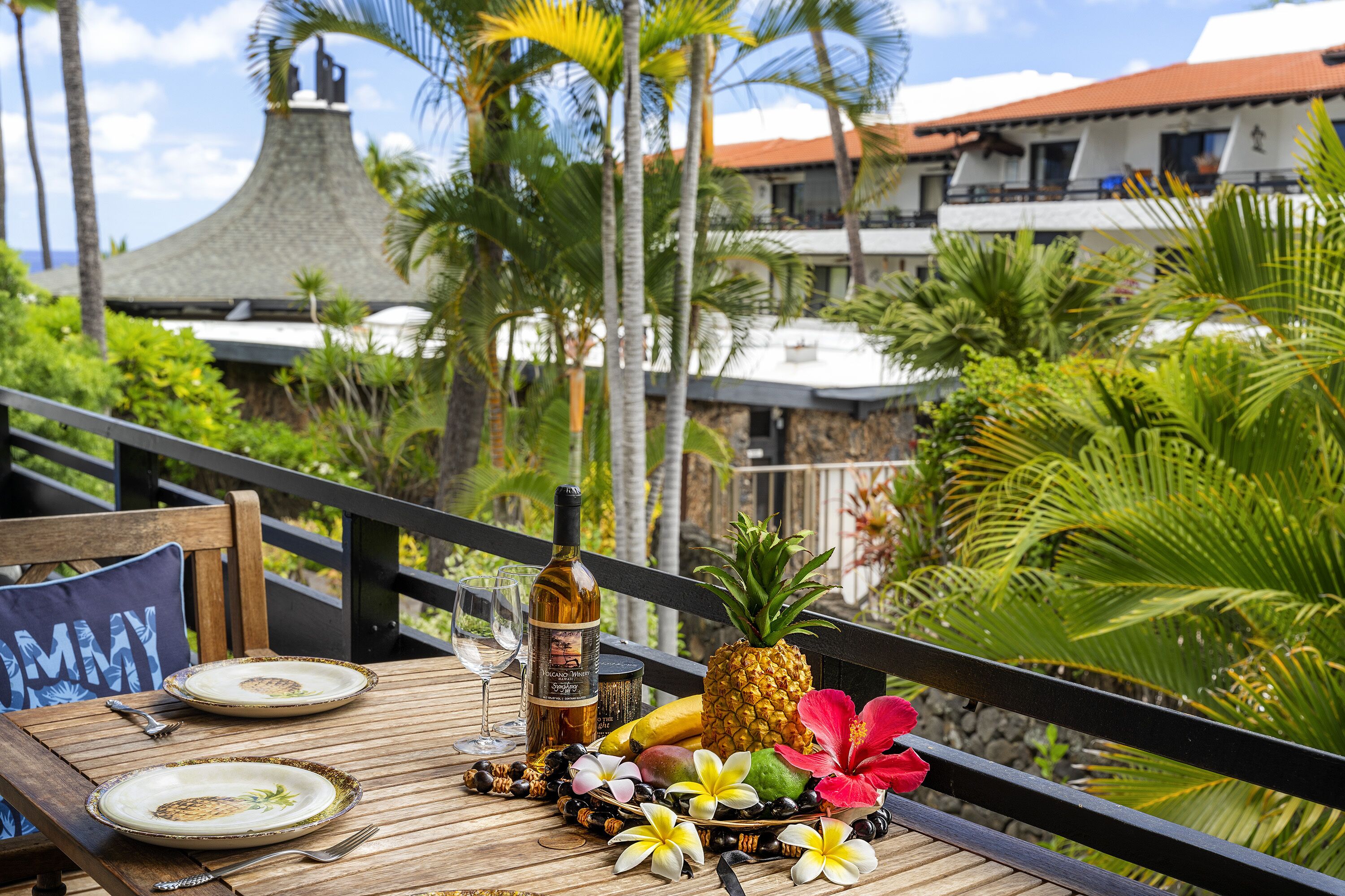 A wooden table with plates, a bottle, a pineapple, and tropical flowers on a balcony with a view of a building and palm trees.
