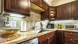 A modern kitchen with dark wood cabinets, granite countertops, stainless steel appliances, and a stone backsplash. - Casa De Emdeko 210- 1/1 DELUXE SIZED UNIT,REMODELED, AC, OCEAN VIEW, ELEVATOR (Kailua Kona)