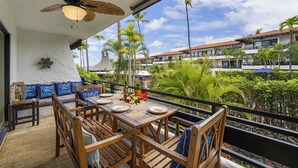 A wooden dining table set with plates, a bottle, and tropical fruits on a balcony with a view of palm trees and a building in the background. - Casa De Emdeko 210- 1/1 DELUXE SIZED UNIT,REMODELED, AC, OCEAN VIEW, ELEVATOR (Kailua Kona)
