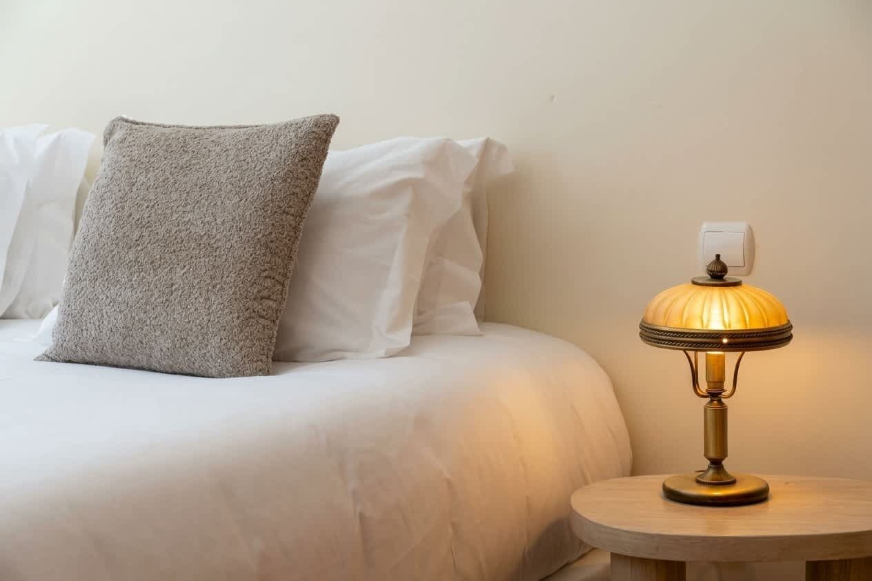 Una cama con ropa de cama blanca y almohada gris, una mesita de noche redonda de madera con una lámpara de mesa y un enchufe.