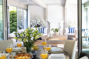 A modern living room with a sofa set, a dining table with breakfast items, and a view of greenery through large windows.
