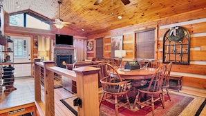 Interior acogedor de una cabaña con techo de madera, chimenea, comedor con sillas de madera y un ventanal grande.
