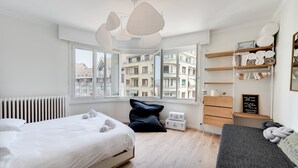 Un dormitorio moderno con cama, ventana con vista a edificios, estante con toallas y cómoda.