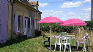 A rustic stone building with purple shutters, a small patio with a table and chairs, and pink umbrellas.