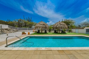 A swimming pool with a white fence, two thatched-roof structures, and a clear blue sky. - 3BR Villa w/ Private Pool near Palm Beach (Noord)