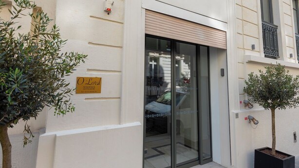 Entrada de hotel com fachada moderna, porta de vidro e uma pequena árvore em vaso.